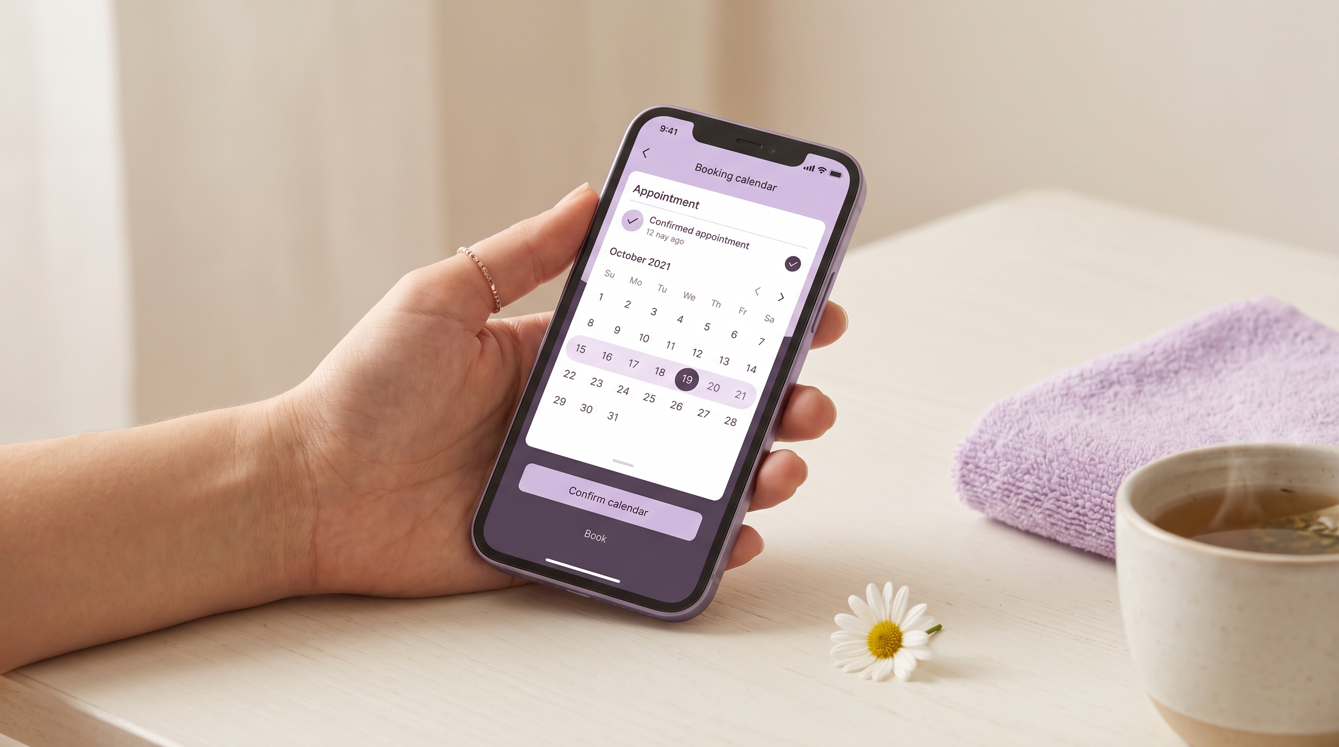 A hand holding a smartphone showing a tidy lavender booking calendar with the next appointment confirmed, on a warm off-white desk with a single daisy bloom